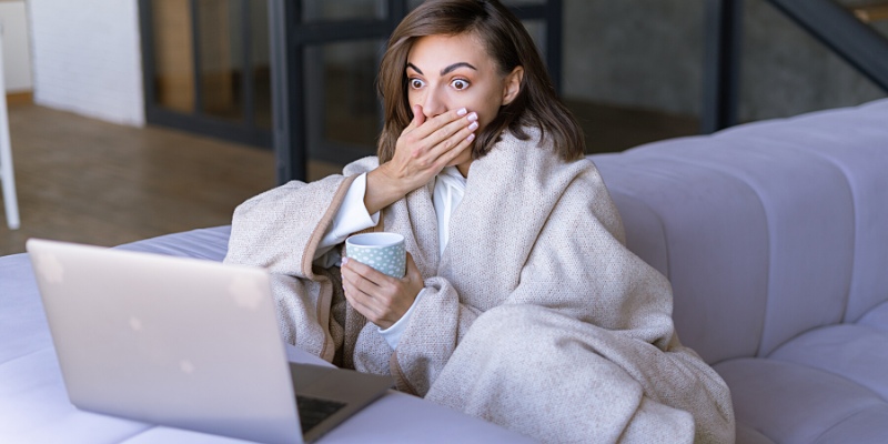 Giovane donna a casa sul divano in inverno sotto un'accogliente coperta con un laptop che guarda scioccata lo schermo per aver perso al casinò di Bwin
